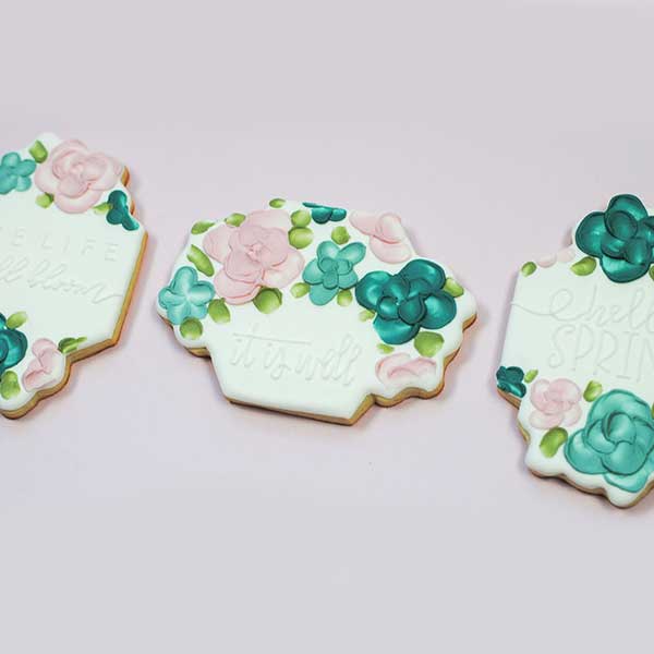 3 designs of floral plaque cookies flooded with a white royal icing base. medium peak royal icing color in vibrant colors and applied with a palette knife to create flowers. White on white spring quote written "it is well", "hello spring" and "live life in full bloom".