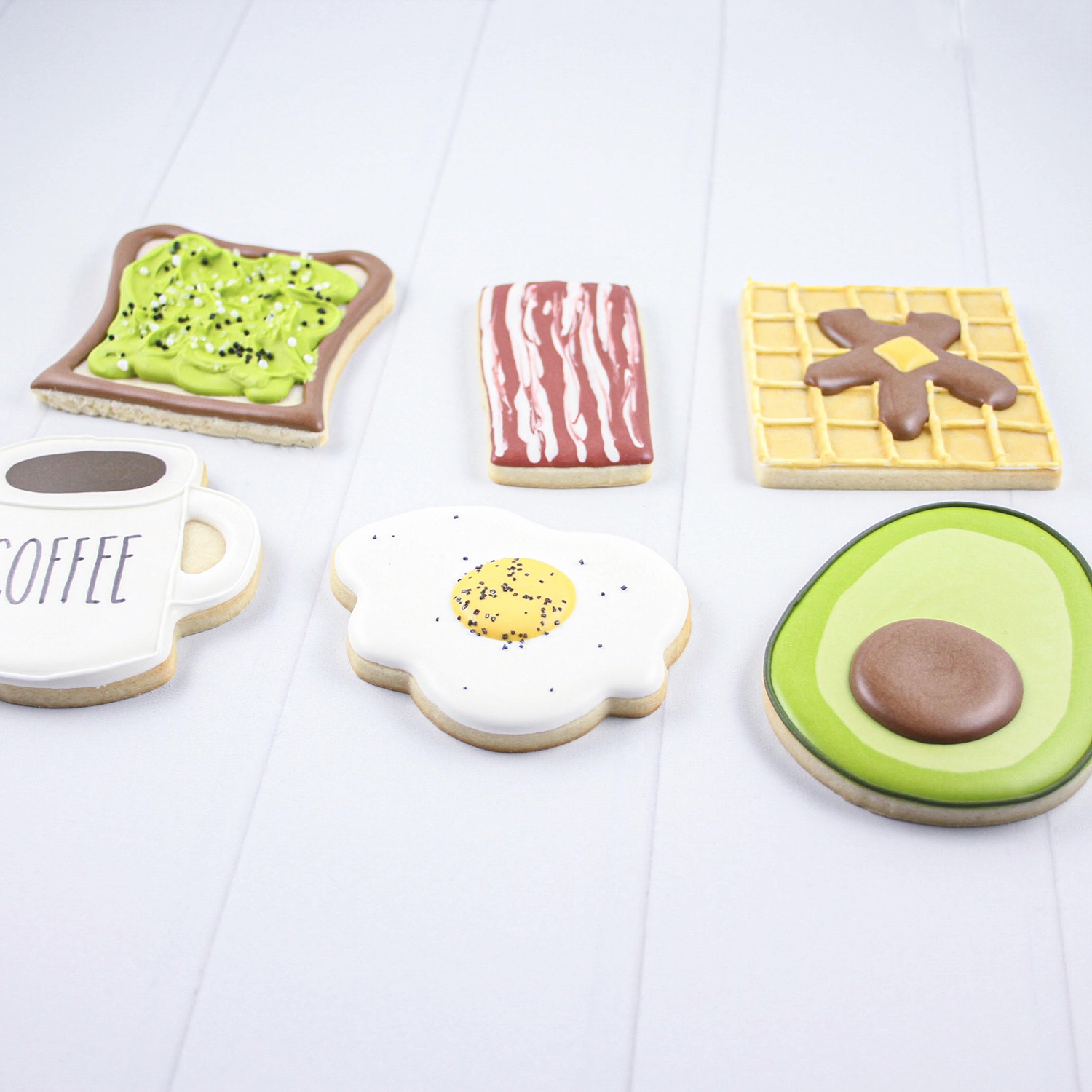 Breakfast Foods Cookies include avocado toast, strip of bacon, waffle with butter and syrup, cup of coffee, an over easy egg and half an avocado all decorated in royal icing.