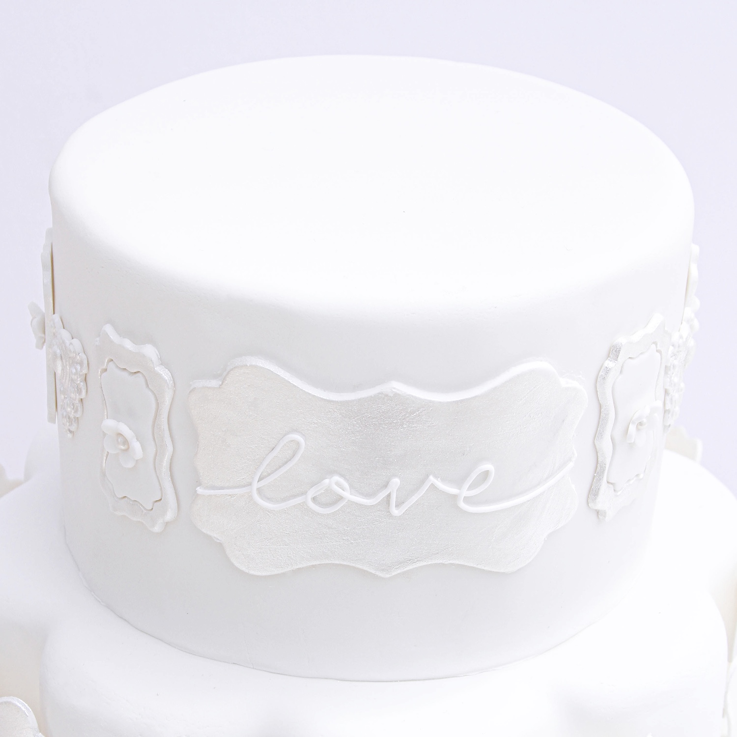 Top tier of cake covered in fondant with nora frame fondant cutouts encircling the side of the cake, covered in luster pearl dust and love hand piped in royal icing.