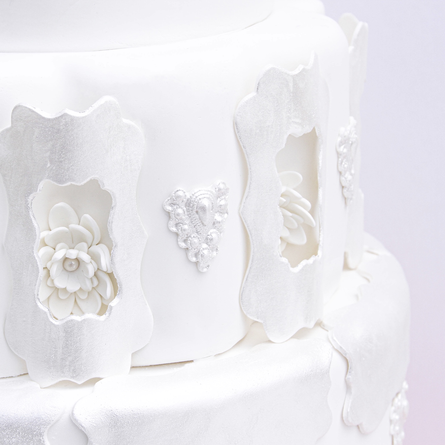 Middle tier of fondant wedding cake with nora frame cutouts which are curved windows framing fondant flowers. There is a fondant pendant between every other curved window.