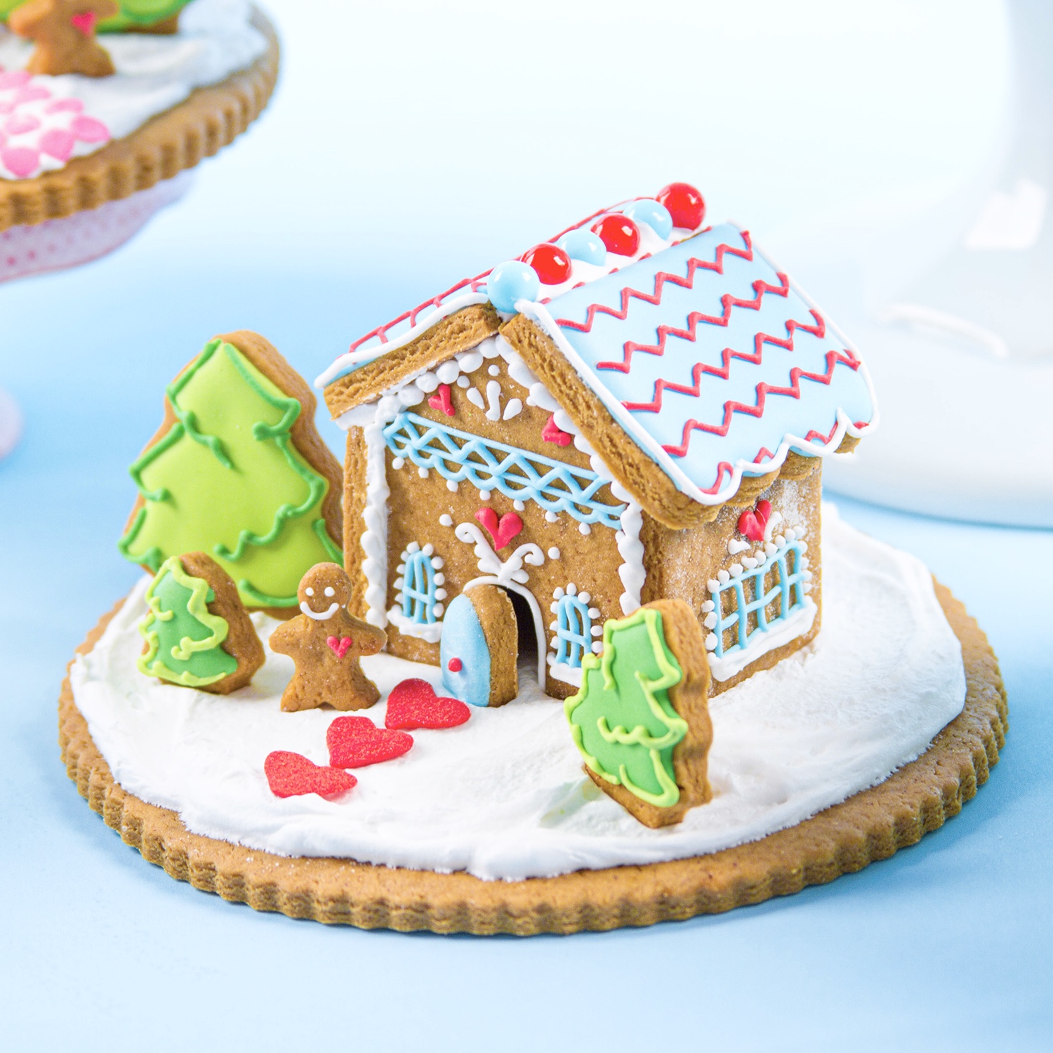 Mini Edible Gingerbread House decorated in royal icing sitting on a snow covered scallop cookie with a candy daisy path, sixlets line the roof top, decorated trees and gingerbread boy cookie make the scene.