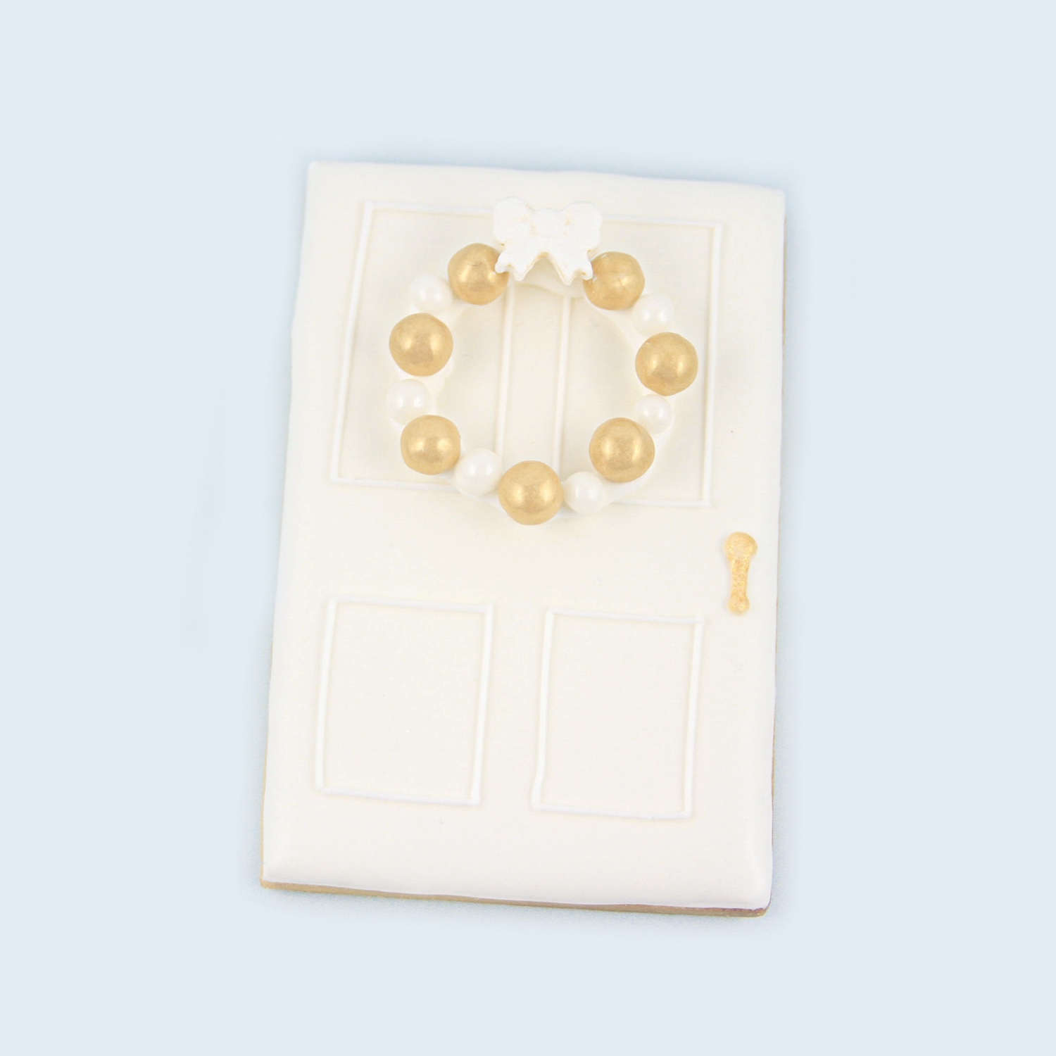 White front door royal icing cookie with a winter wreath made of white and gold sugar pearls and a candy bow. Gold painted doorn knob