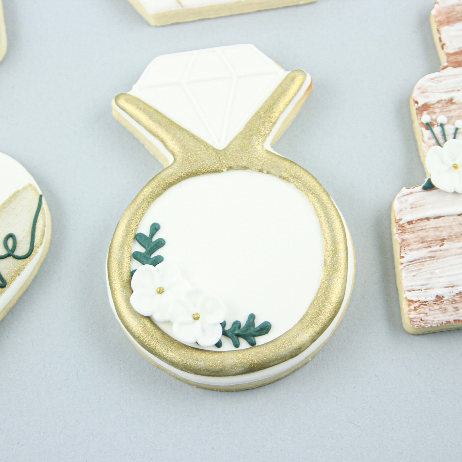 Engagement diamond ring cookie decorated white a white background, gold ring and adorned with white royal icing floral accents.