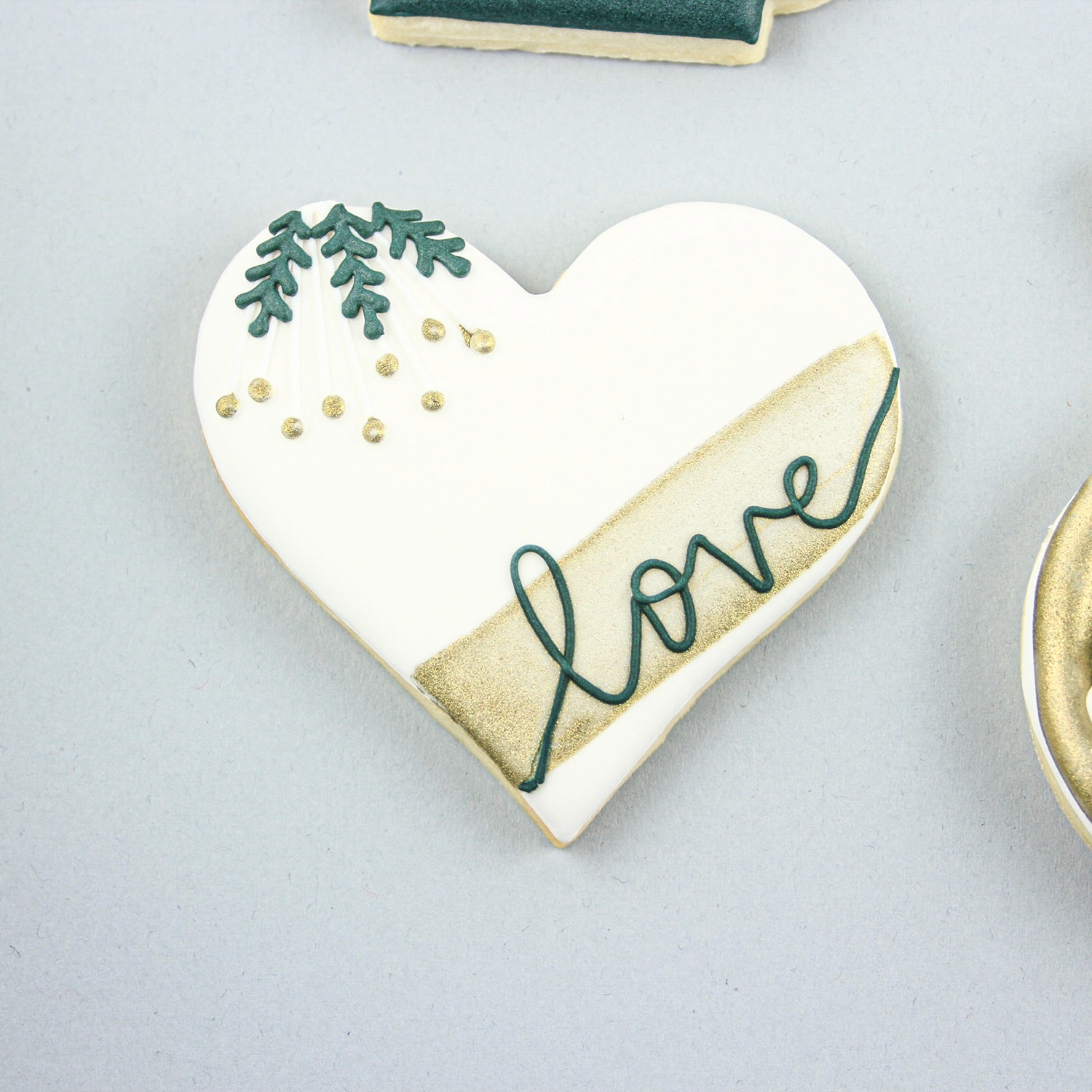 Heart shaped cookie decorated in white royal icing, accented with green royal icing piping leaves, adorned with white royal icing flowers and painted accents in gold and love written in green.