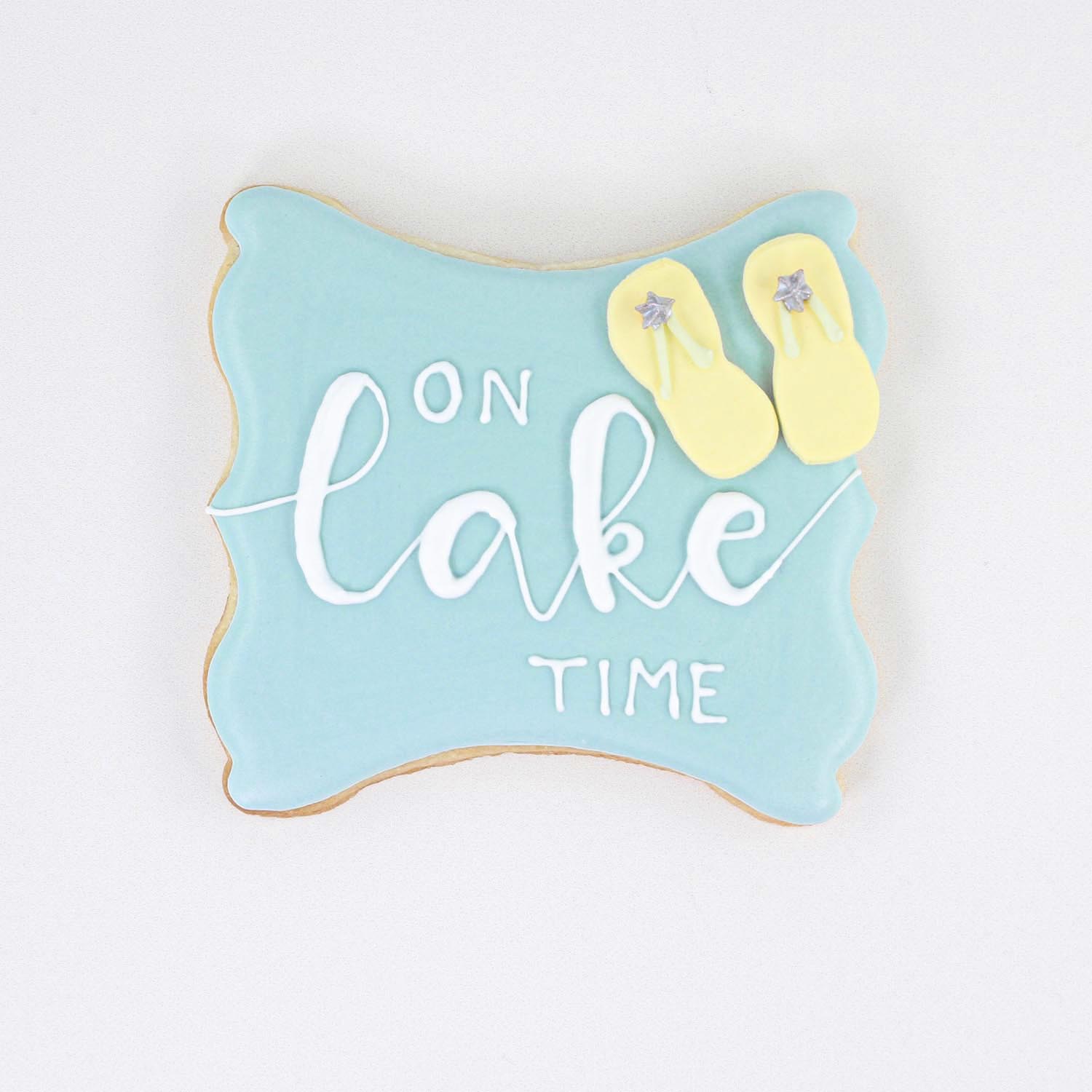 "On Lake Time" plaque cookie decorted in blue and white royal icing and adorned with yellow edible flip flops.