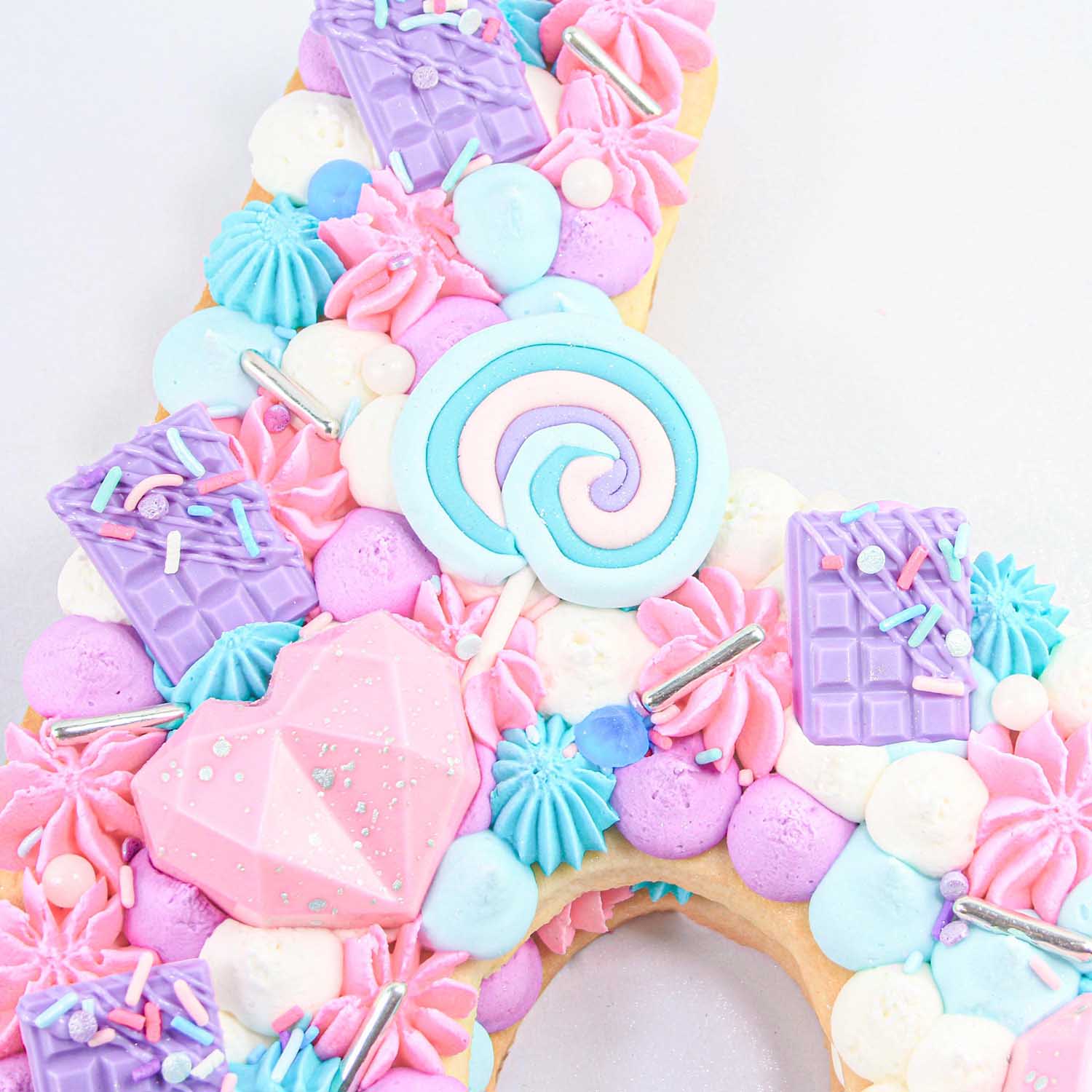 Sweet 16 Number Cookie Cake decorated  in shades of pink, purple and blue dots of buttercream using round, open and closed star piping tips. Buttercream is adorned in molded chocolate bars and faceted hearts and sprinkles and fondant swirled lollipops.