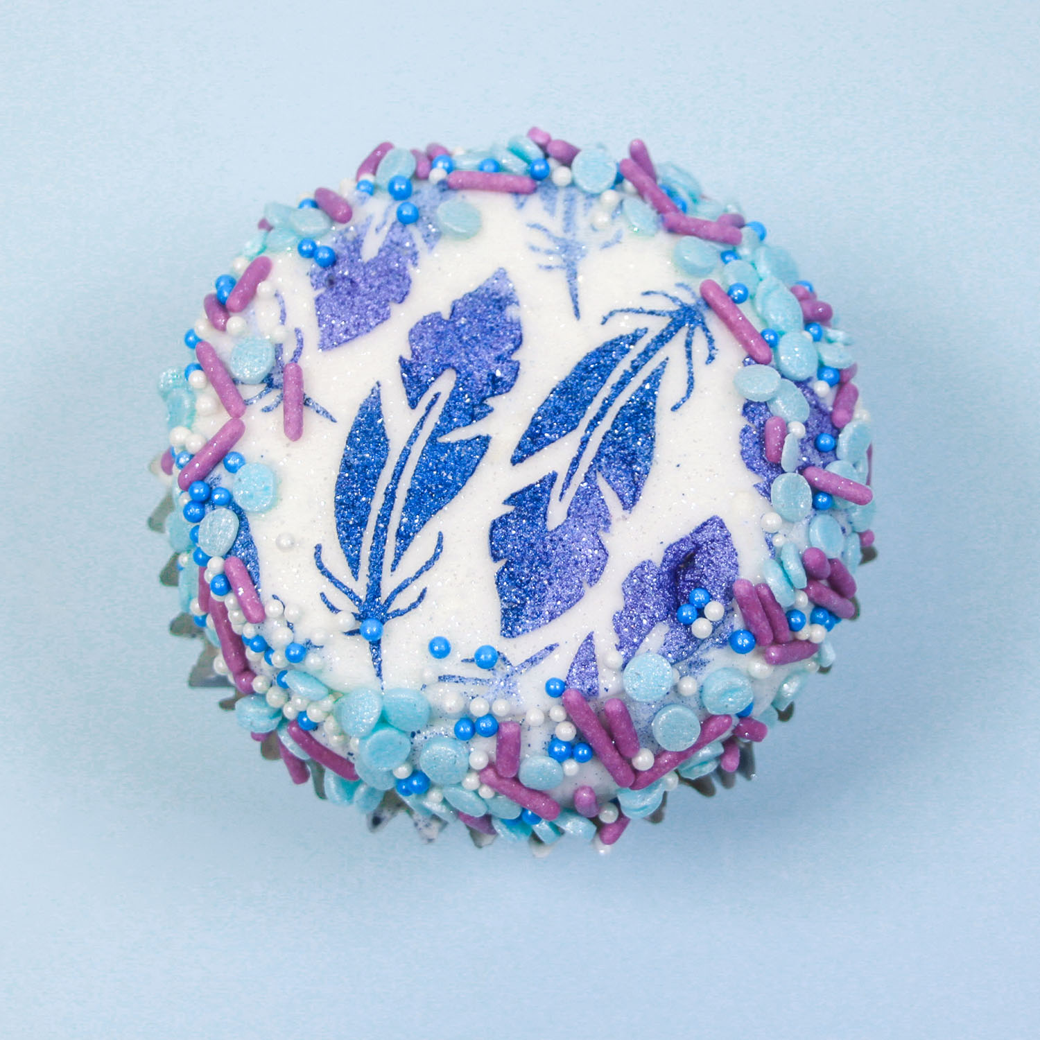 Buttercream feather stenciled cupcake brushed with blue and periwinkle luster dusts and sides of buttercream are dipped in a complimenty sprinkl mix of jimmies, confetti and nonpareils.